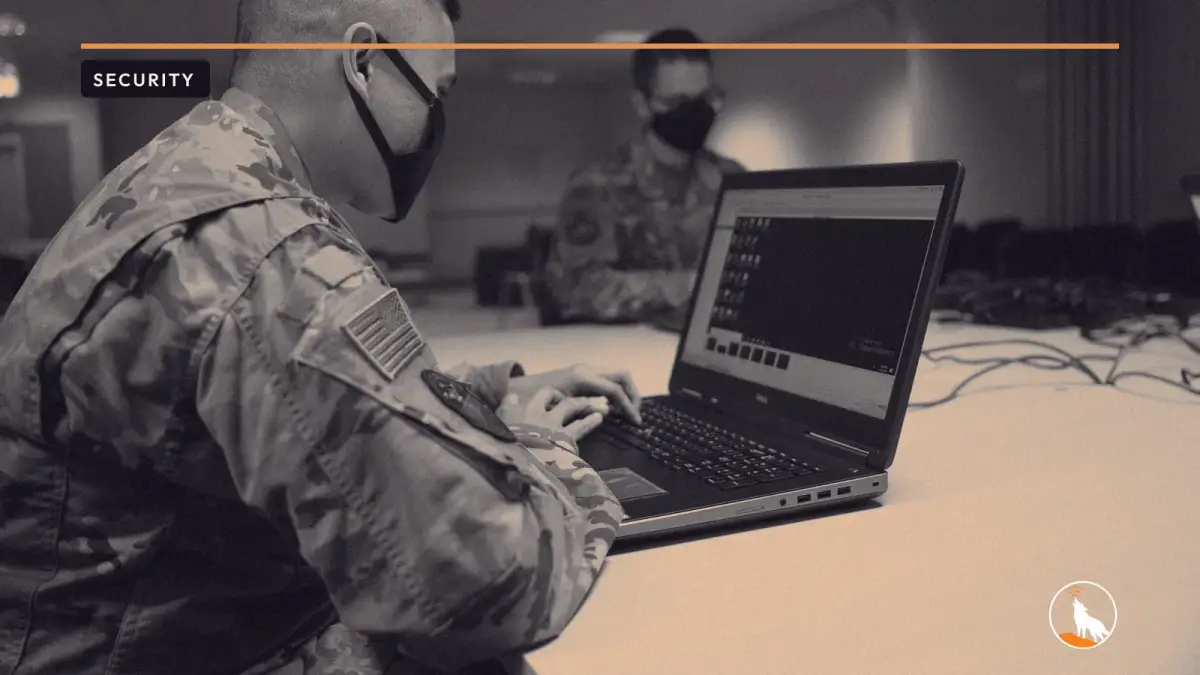 A defensive cyber operator at a workstation, graded navy and orange, with the wolf mark.