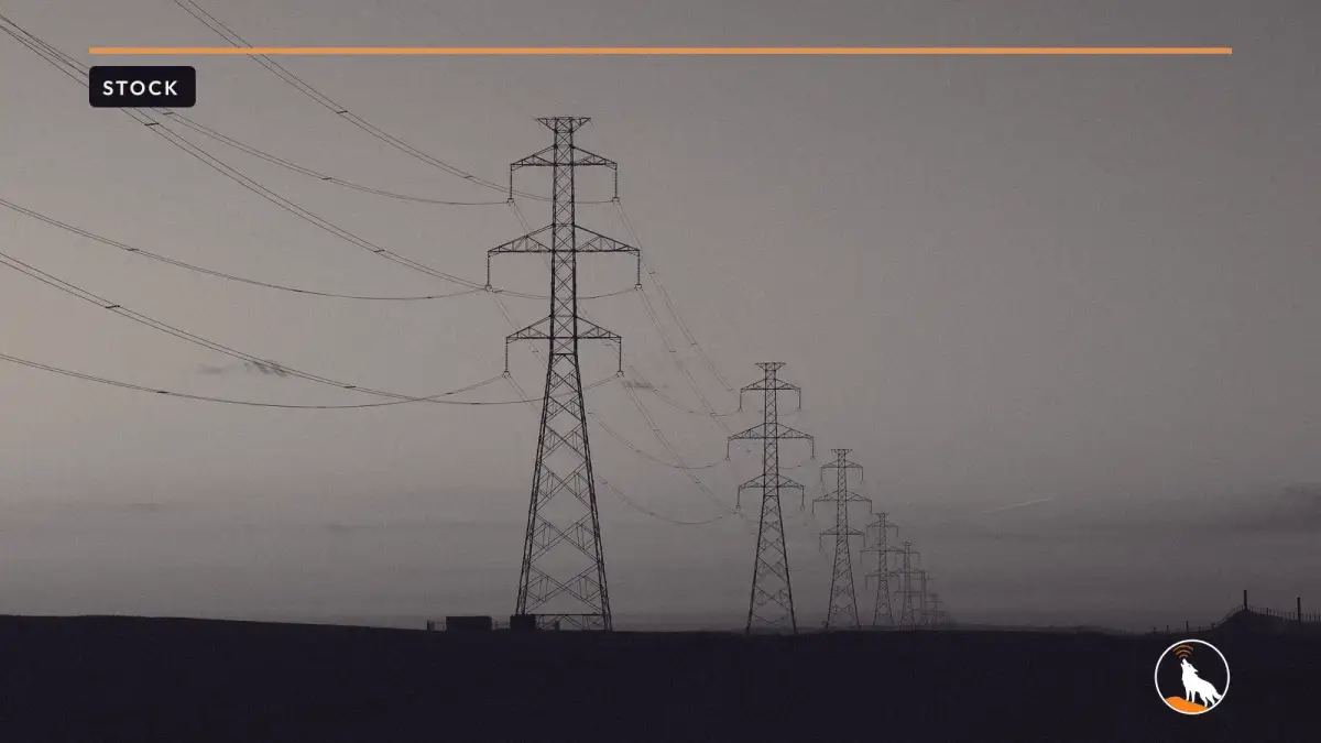 A row of steel transmission towers and power lines silhouetted against an orange sunset fading to blue.