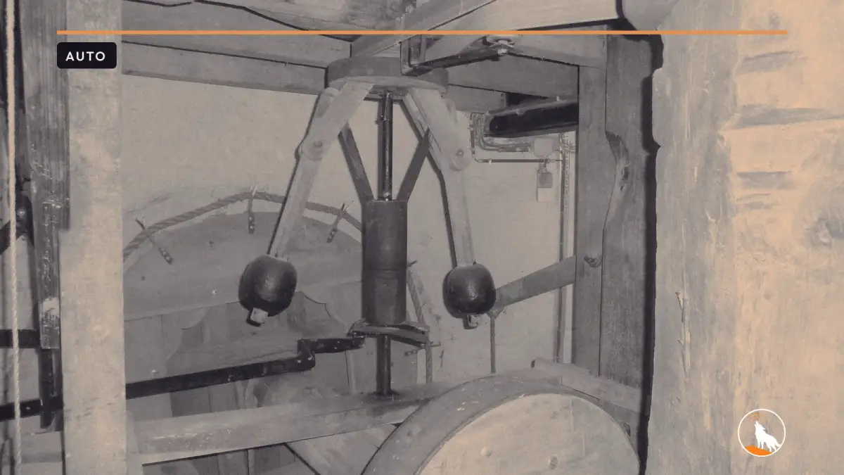 An old centrifugal governor inside a windmill, with two wooden arms holding black iron weights around a central shaft, surrounded by worn timber beams and a wooden wheel.