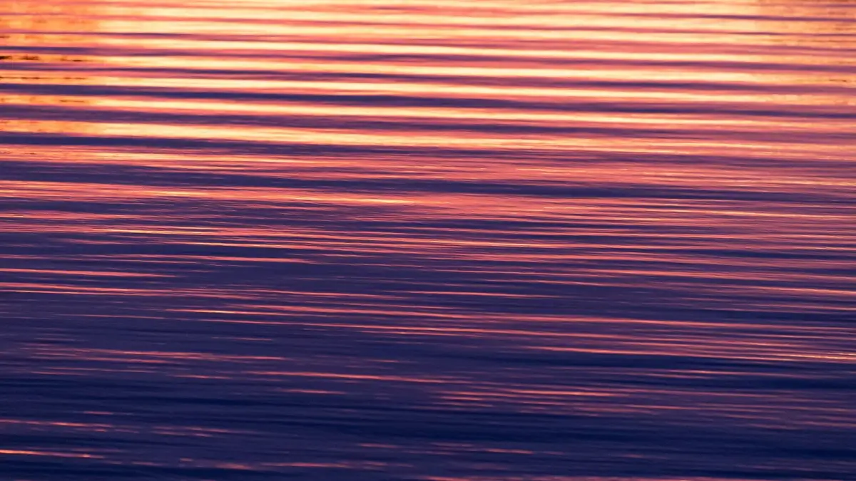 Close-up of rippled lake water reflecting a sunset in horizontal bands of orange, pink and deep purple.