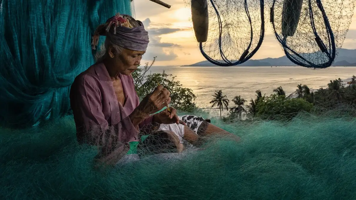 An elderly woman in a headscarf mends a green fishing net piled around her, with hand nets hanging overhead and the sea glowing at sunset behind her.