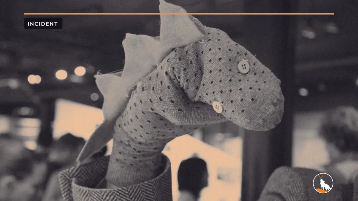 A grey polka-dot sock puppet with button eyes and a spiky blue felt crest, held up on a hand in a tweed sleeve against a blurred crowd.