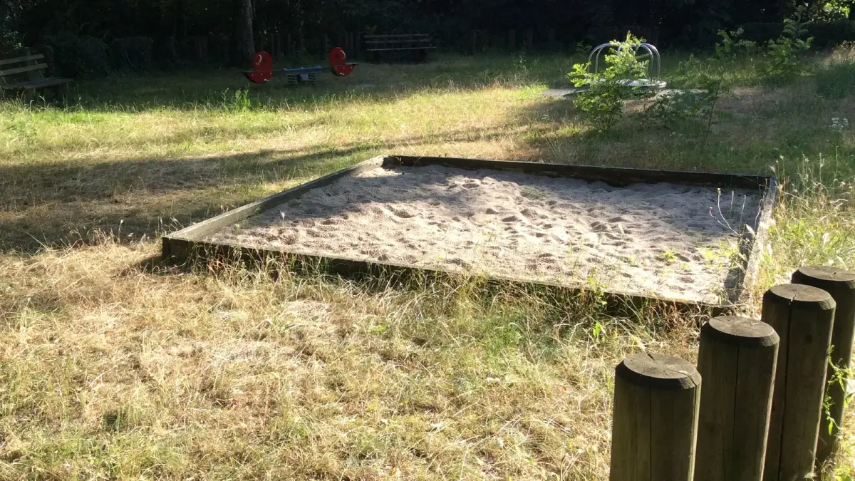 An empty wooden sandbox in long, sunlit grass, with four wooden posts in the foreground and a seesaw and bench in front of dark trees behind it.