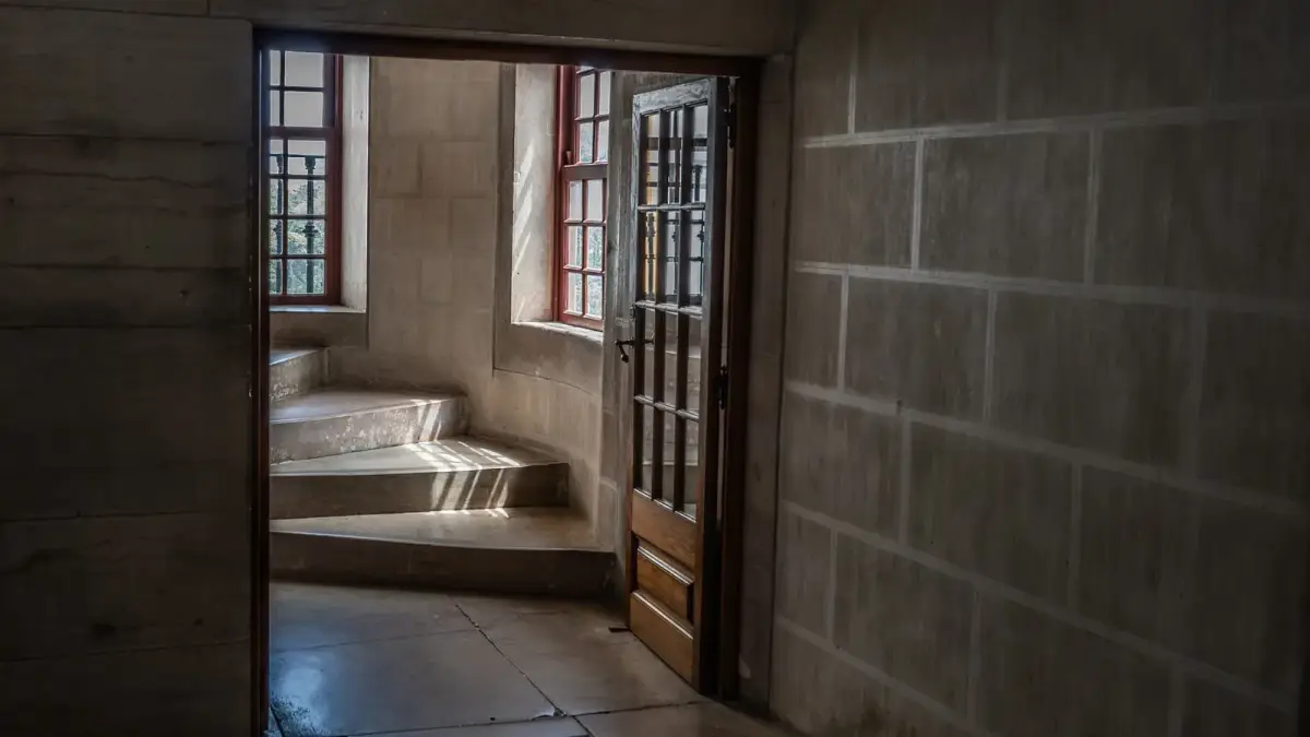Sunlight from two red-framed windows casts striped light across a curved stone staircase, seen through an open wooden door from a dim stone hallway.