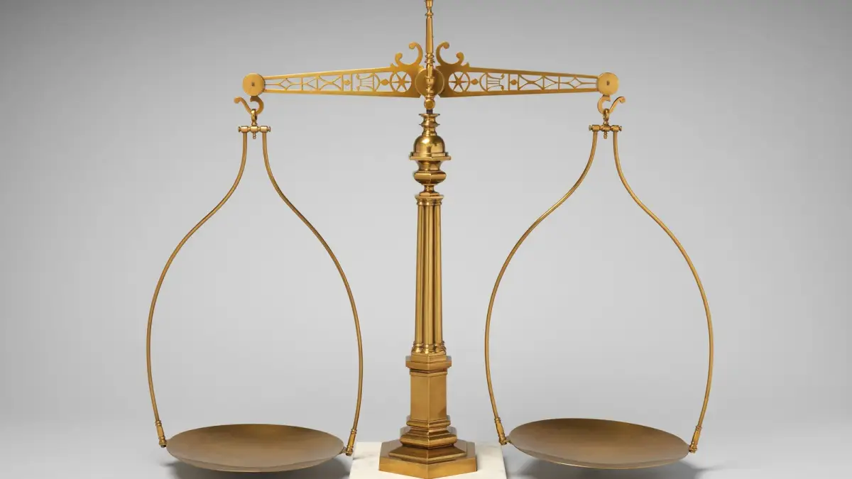 An antique brass balance scale with two empty round pans hanging from an ornate beam, on a white marble base against a gray backdrop.